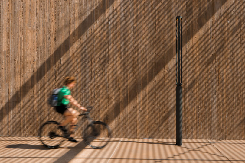 A person wearing a green shirt and a backpack rides a bicycle past a wooden wall with striped shadows and a black pol light.