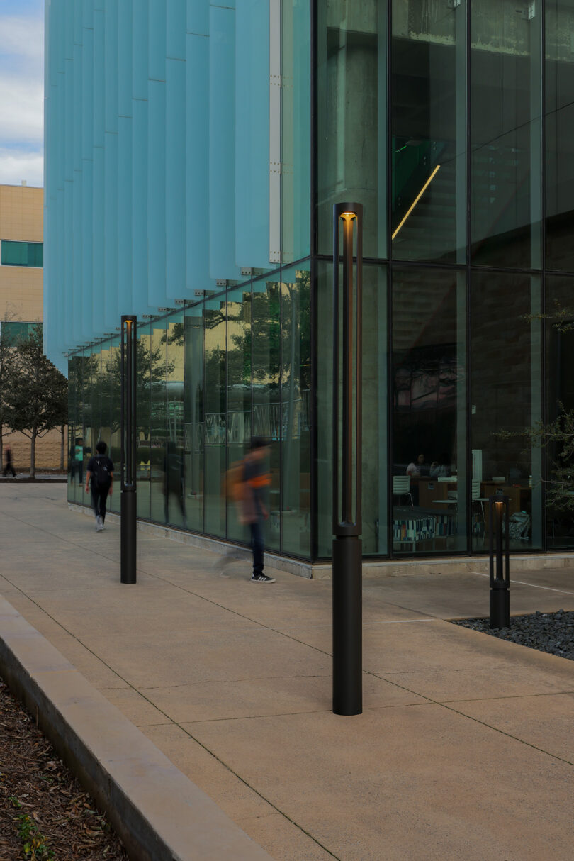People walk by modern glass buildings. The sidewalk is lined with tall, slim black light posts.