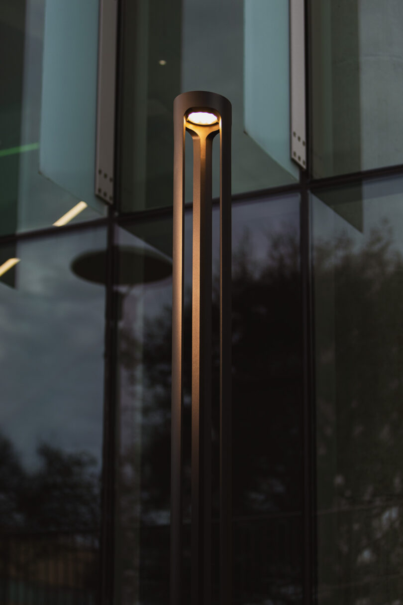 Tall, modern outdoor lamp post with a cylindrical light fixture, illuminated against a reflective glass building background.