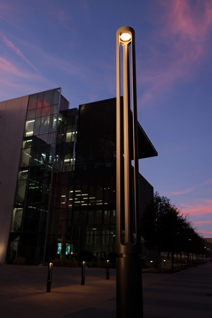 A modern streetlight illuminates a pathway in front of a glass-front building at dusk, with a purple and pink sky in the background.