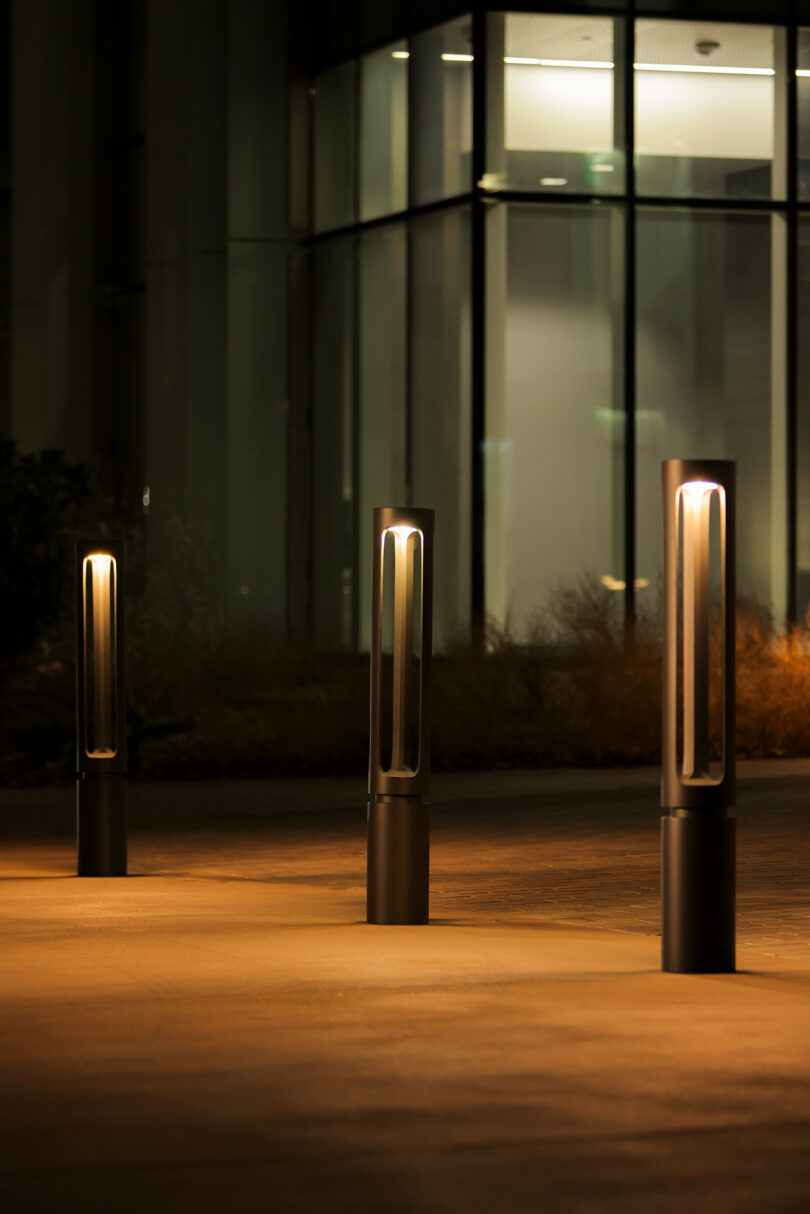 Three modern, cylindrical outdoor lights illuminate a pathway in front of a large glass building at night.
