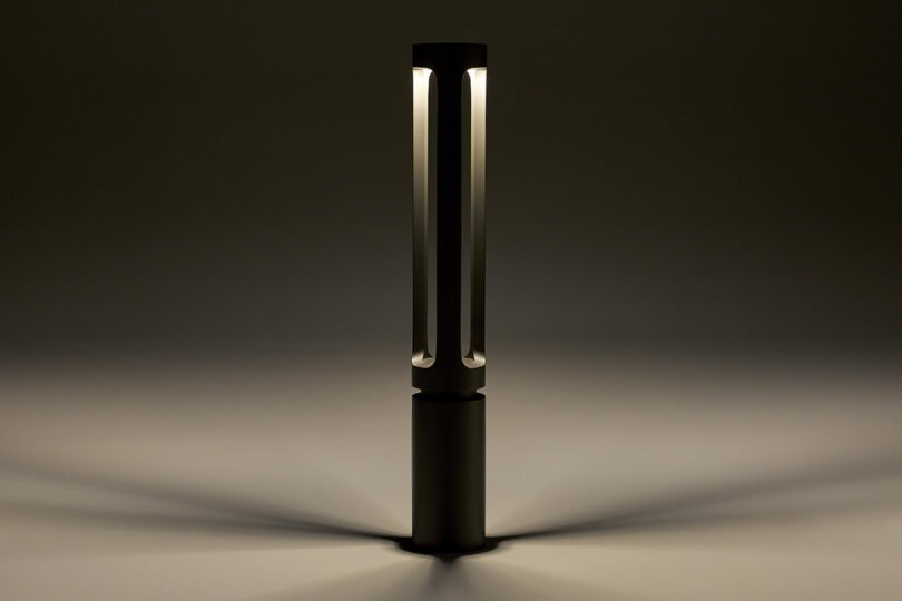 A modern lamp emits soft light in a dark room, casting shadows in a radial pattern. The lamp features a slim, cylindrical design.