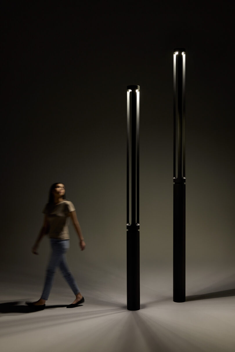 A person walks past two tall, modern, black lamps in a dimly lit setting.