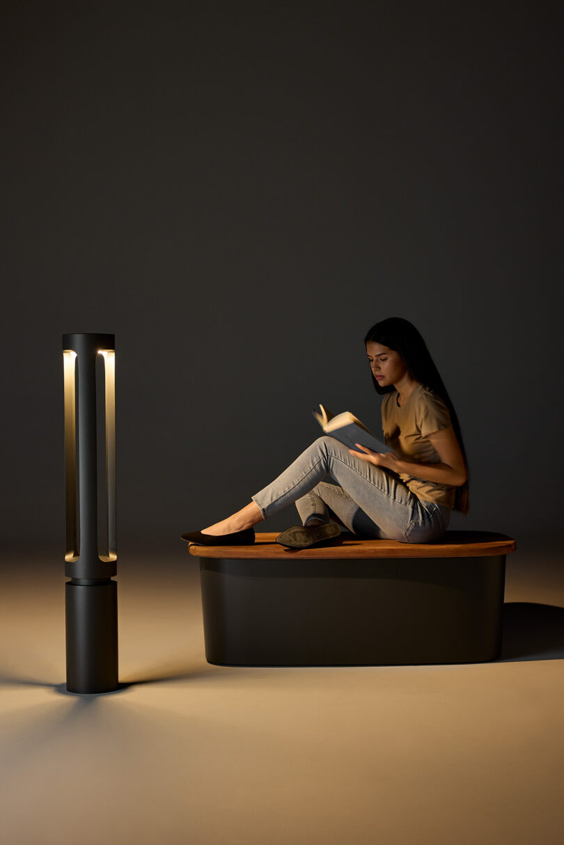 A woman with long hair sits reading a book in a dimly lit room. A tall, cylindrical lamp illuminates the scene.