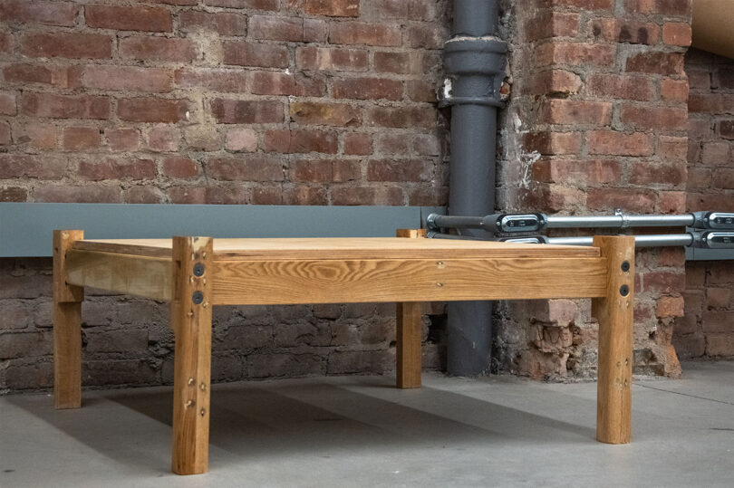 A square wooden coffee table with thick legs placed against a brick wall with a grey pipe running vertically beside it.