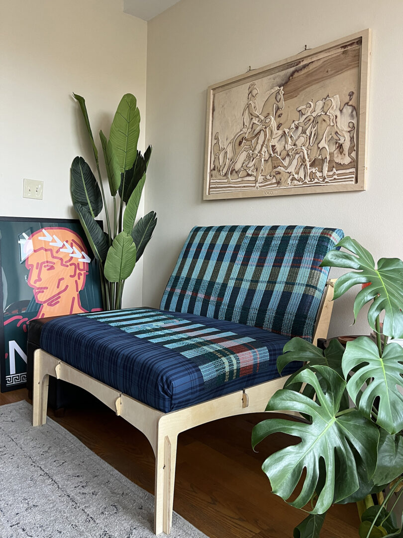 A modern daybed with a blue and green plaid cushion. There's art above it, a potted plant on each side, and a colorful poster leaning against the wall.