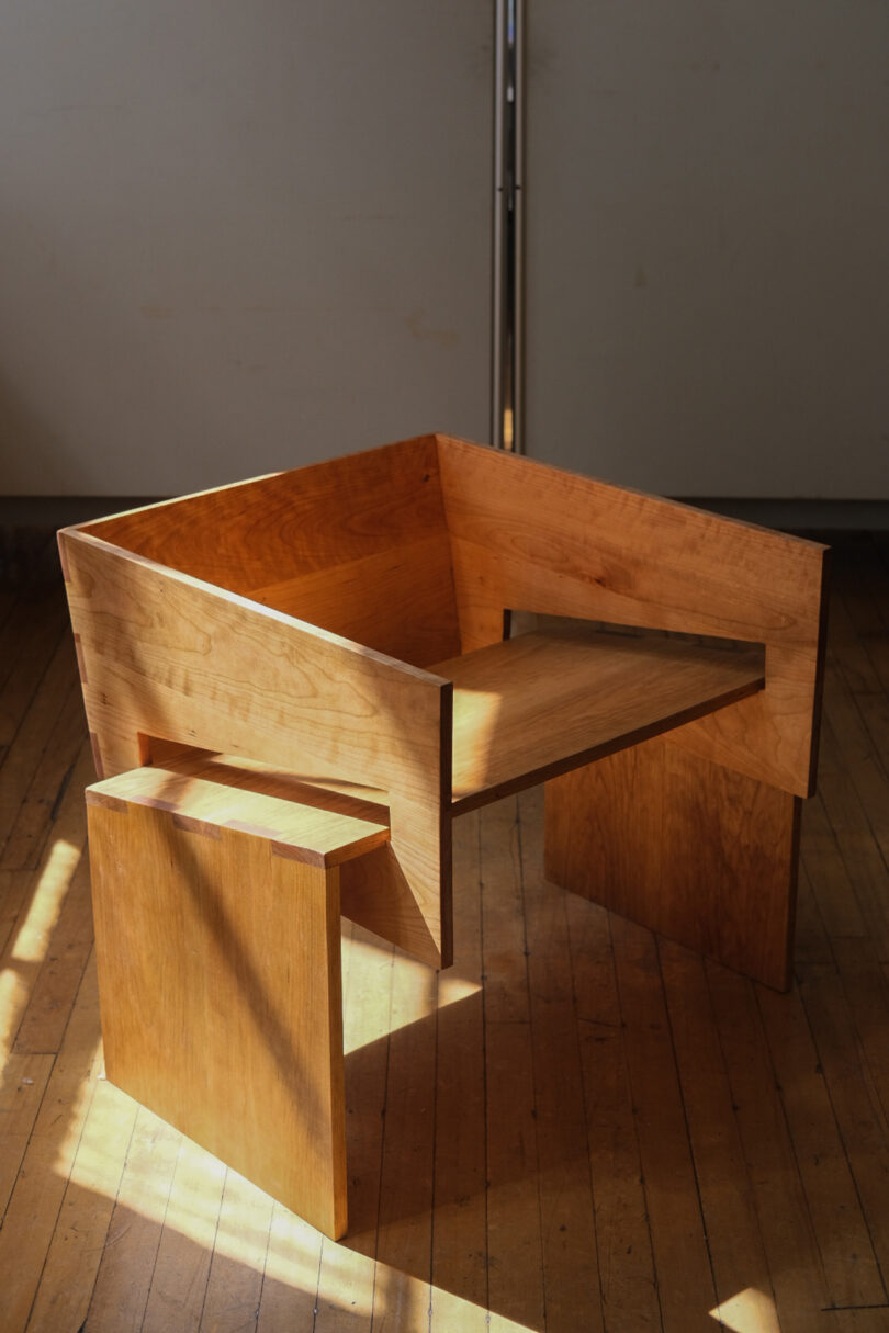 A wooden chair with a minimalist design sits on a hardwood floor. Sunlight creates a pattern of light and shadows on the chair and floor. The chair has a square back and flat armrests.