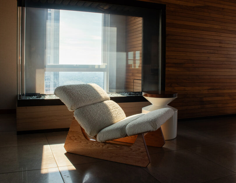 A modern lounge chair with a wooden frame and white cushioned seat is placed by a large window, with sunlight streaming in and accenting the wood-paneled wall in the background.