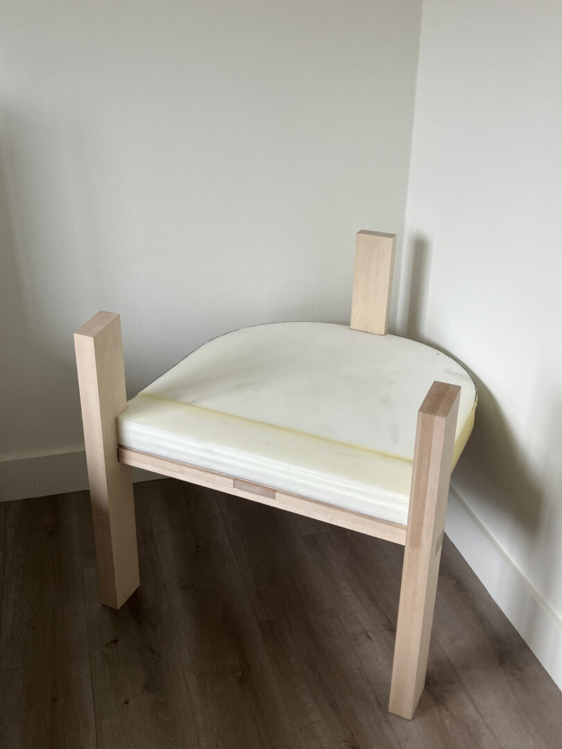 A wooden chair with light-colored legs on a hardwood floor with a white, semi-cushioned seat leaning against a wall.