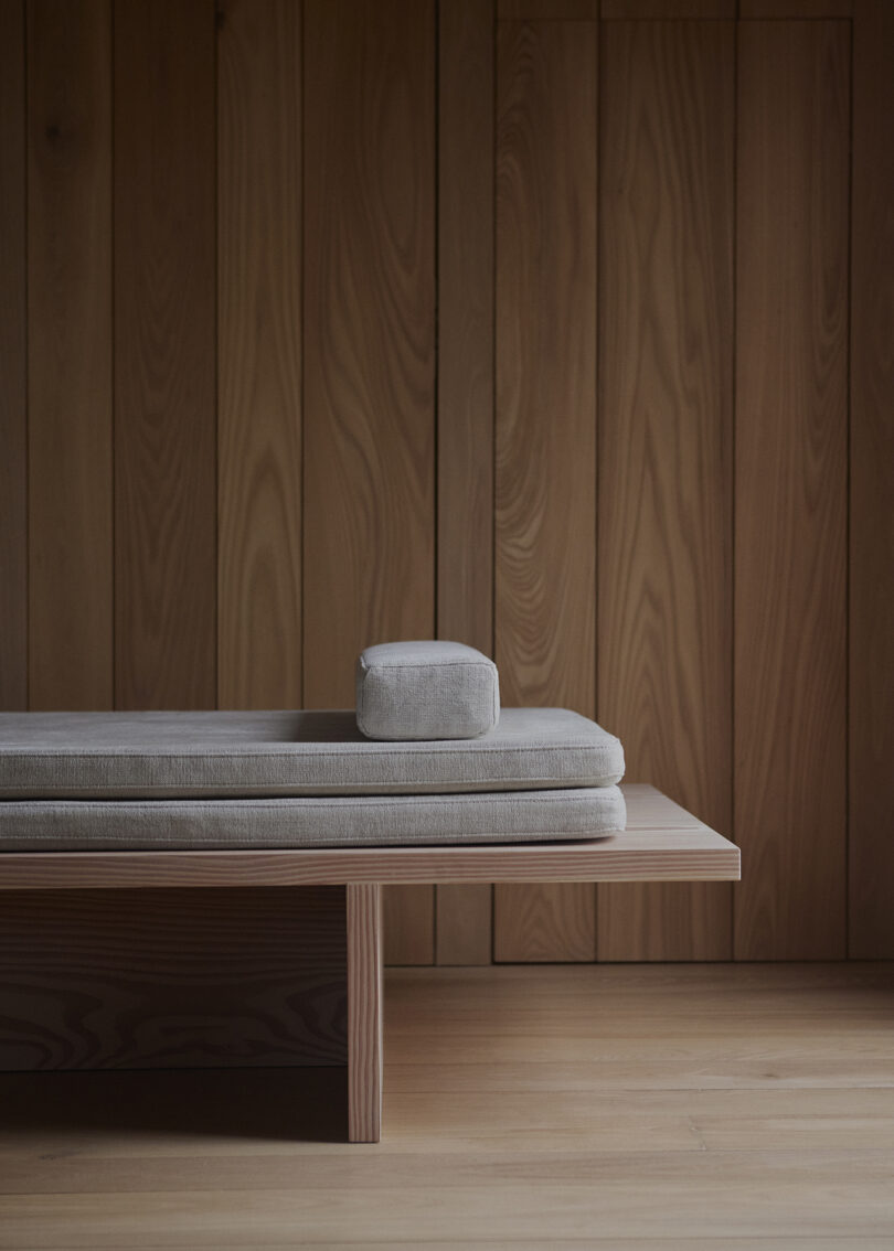 A minimalist wooden daybed with a light gray cushion and a small headrest in front of wood-paneled walls.