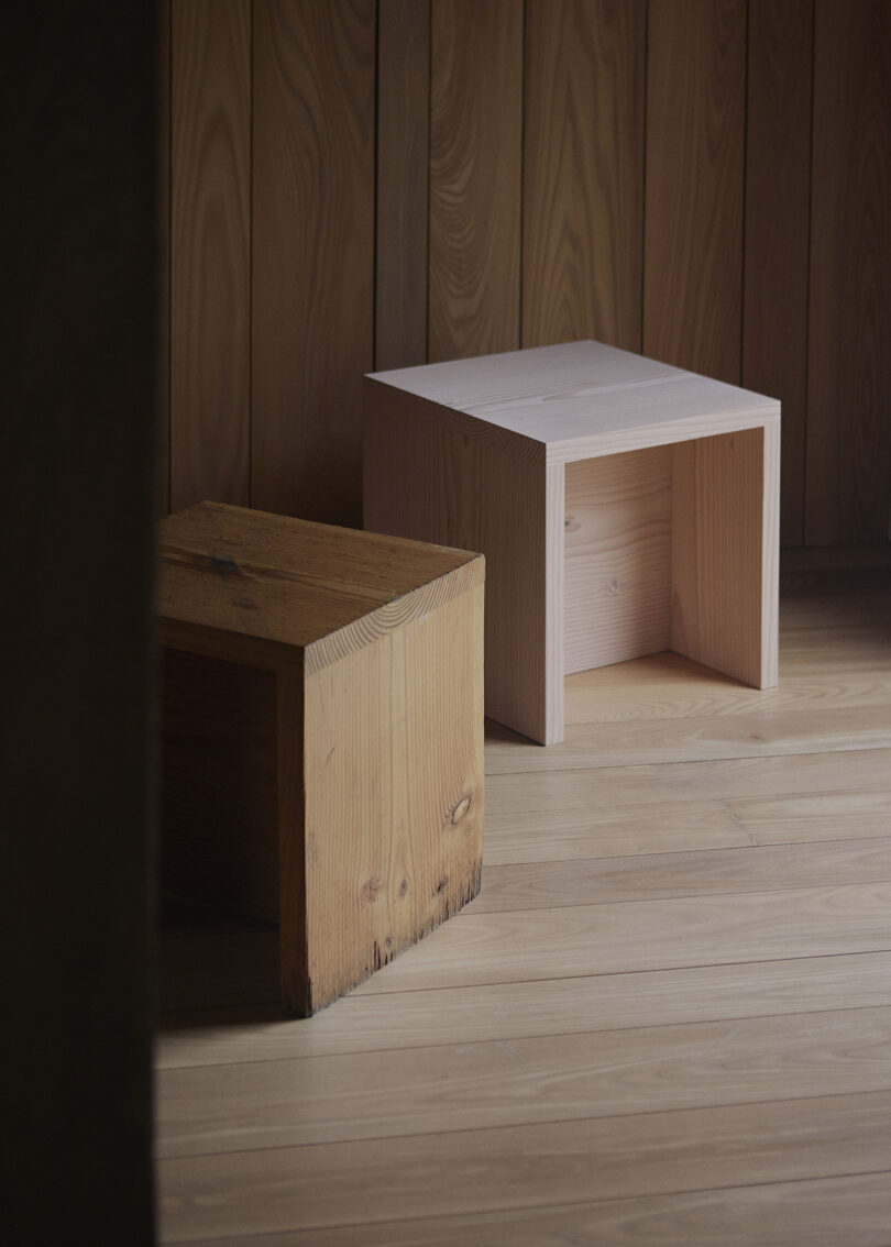 Two wooden stools, one lighter and one darker, placed on a wooden floor against a wooden wall background.