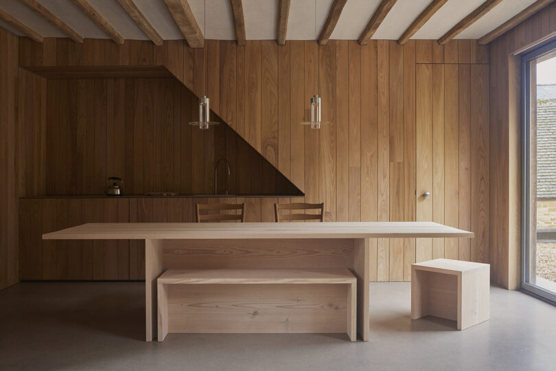 Minimalist wooden kitchen with a long dining table, a bench, and a cubic stool under exposed ceiling beams. A teapot sits by the sink under angled lighting fixtures.