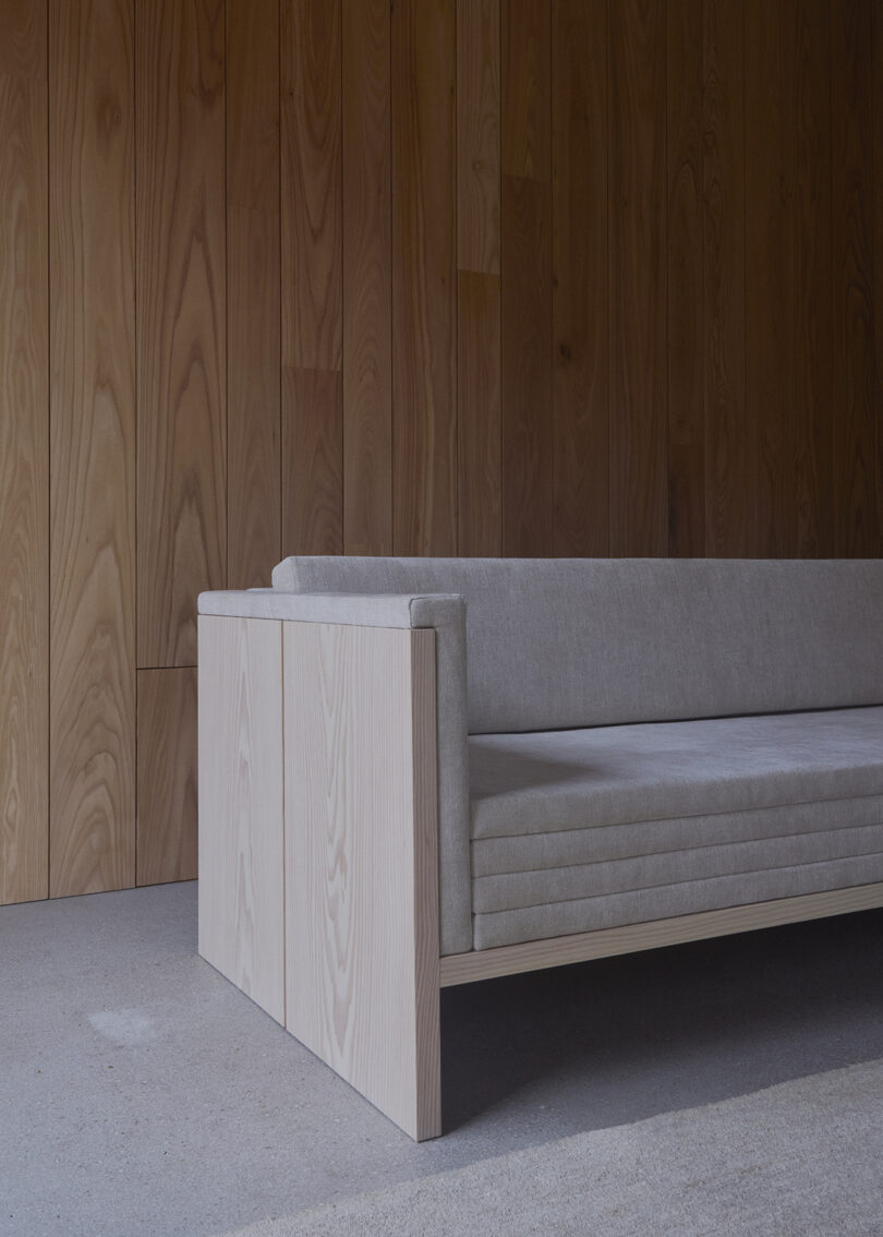 A minimalist beige sofa with clean lines is placed against a wooden-paneled wall and a concrete floor.