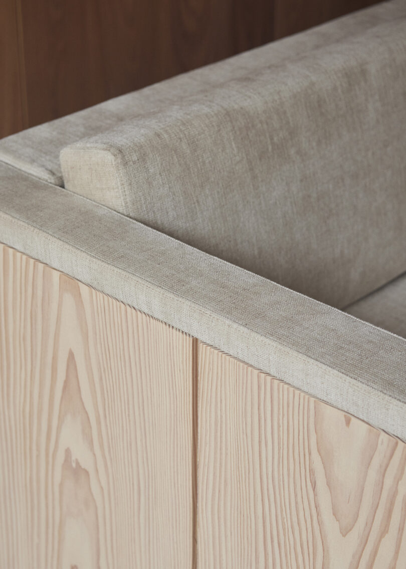 A close-up view of a beige fabric sofa with wooden armrests and back paneling, showcasing the texture and materials used in the furniture design.