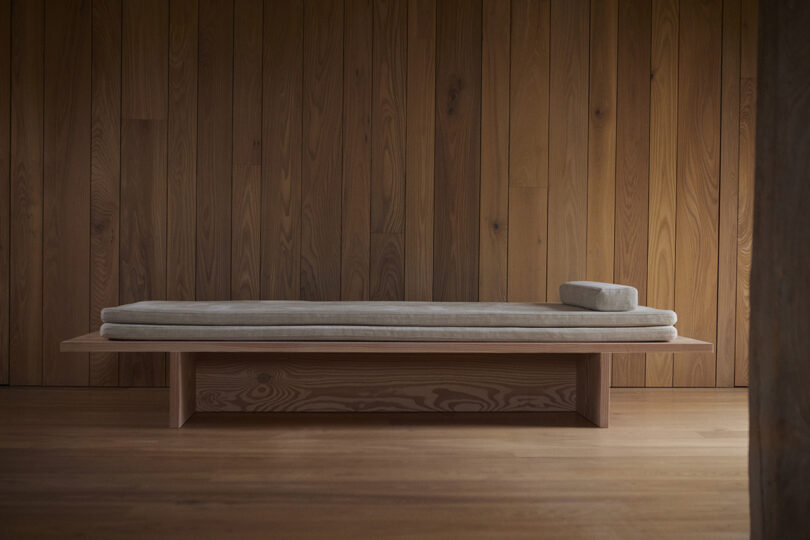 A minimalist wooden daybed with a light gray cushion and a small headrest in front of wood-paneled walls.