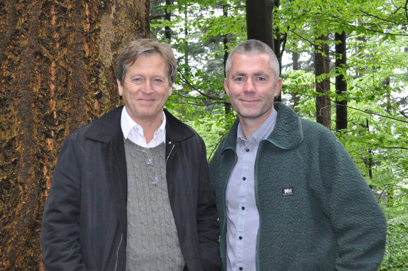 Two men stand side by side in a forested area, one wearing a dark jacket over a sweater, the other in a green fleece jacket. A large tree trunk is visible in the background.