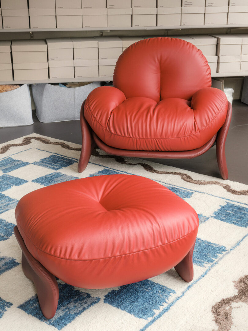 A plush red leather chair with a matching ottoman sit on a white and blue checkered rug in front of shelves filled with beige boxes.