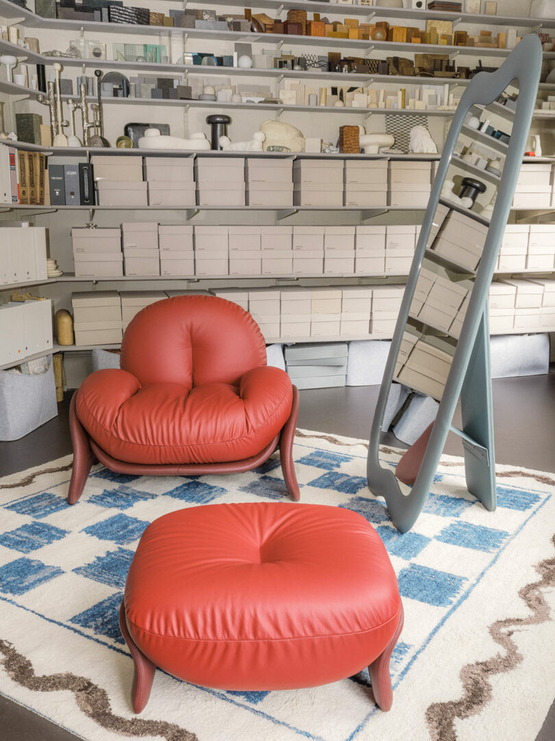 A room with a red cushioned chair and matching ottoman on a blue and white geometric rug, a tall framed mirror, and shelves filled with various items in the background.