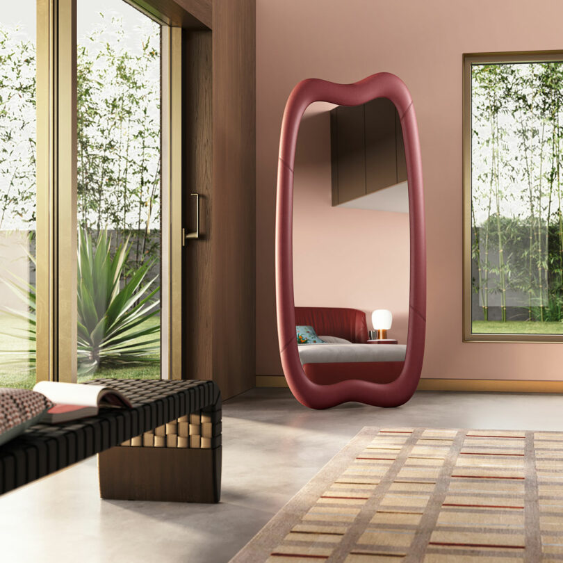 Modern room with large pink-framed mirror, wooden bench, patterned rug, small table with lamp, and large windows showcasing outdoor greenery.