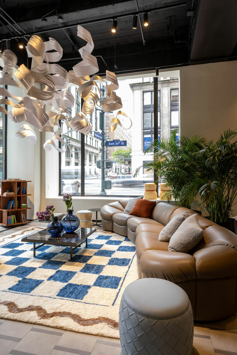 Modern living room with brown sectional sofa, blue and white checkered rug, unique ceiling light fixture, potted plants, and large windows overlooking city street.