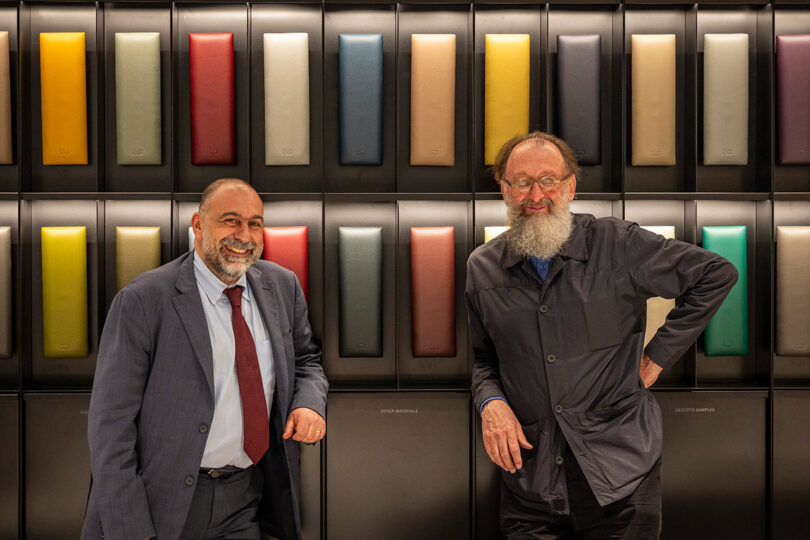 Two men pose in front of a wall displaying a variety of colored materials. One man wears a suit, and the other has a beard and wears a jacket. Both are smiling.