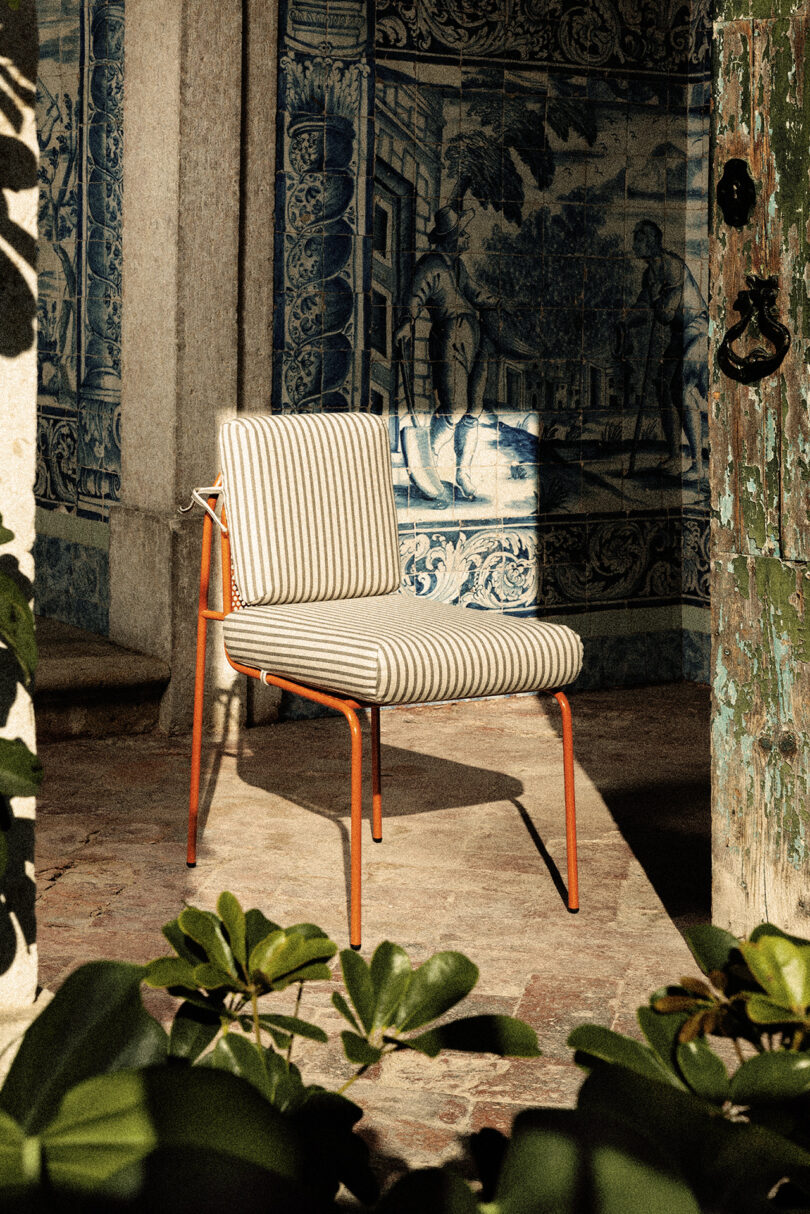 A chair with an orange frame and striped cushion sits in a sunlit area surrounded by greenery.