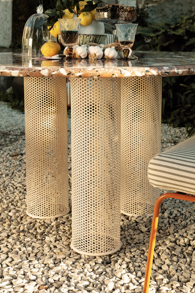 A round table with a mosaic top and three cylindrical, perforated legs. On the table are glasses, a cake stand with fruit, and a glass cover with lemons. Gravel and a striped chair are in the background.