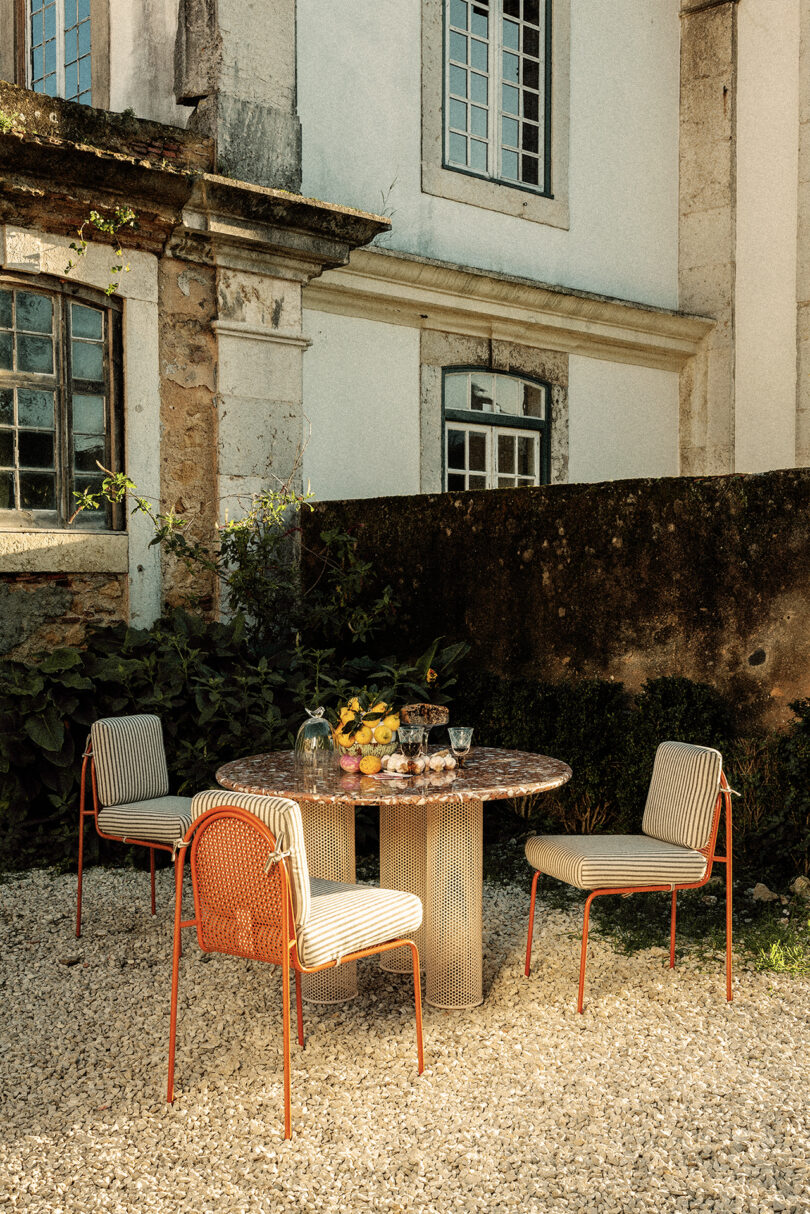 Outdoor dining setup with a round marble table and four striped chairs on a gravel surface in front of an old stone building. A centerpiece of fruit and flowers is placed on the table.