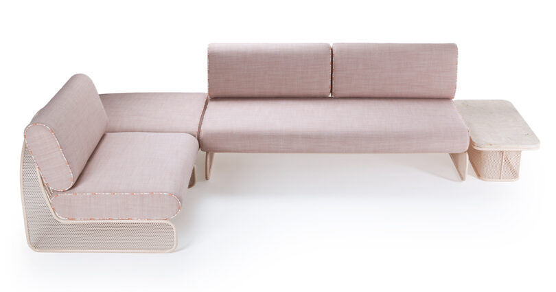 A L-shaped modular sofa with light pink cushions and a built-in side table. The sofa has rounded edges and a perforated base design.