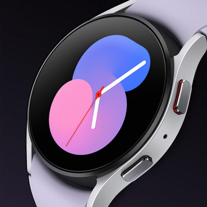 A close-up of a smart watch display showing an abstract design with overlapping pink and blue circles and a white minute and red hour hand. The watch has a metallic bezel and white strap