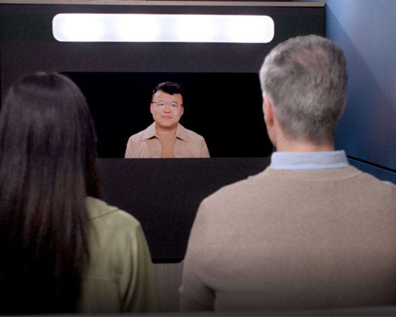 Asian man wearing glasses on a screen being viewed by a woman with long dark hair and a man with salt and pepper hair looking at him illuminated by a long oblong light from above the screen.