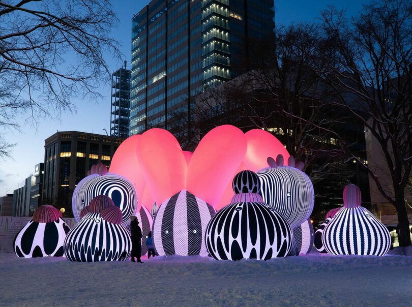 Outdoor art installation with large, abstract, spherical structures in black-and-white patterns and pink lighting, set in a snowy urban area with tall buildings in the background at dusk.