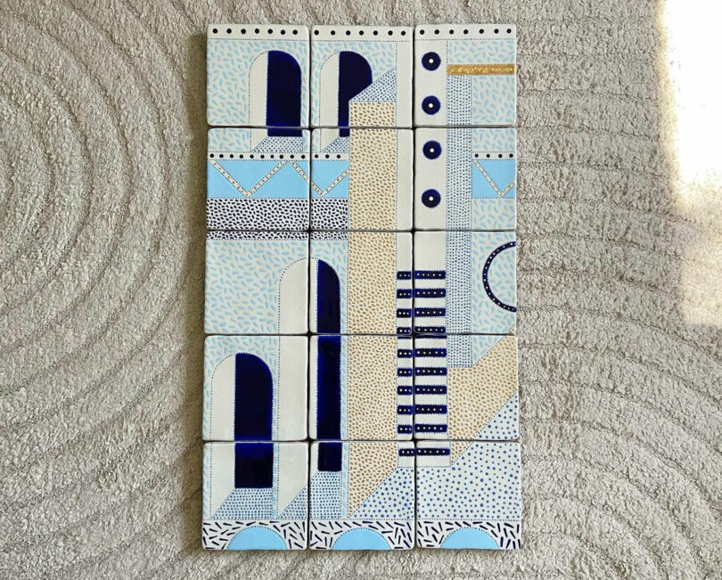 A set of 18 rectangular tiles with a geometric blue, white, and beige pattern arranged in a grid, placed on a textured beige surface.