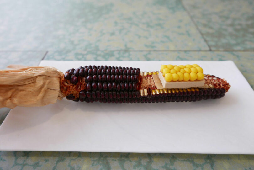 Image of a dark-colored ear of corn on a white rectangular plate, with a small piece of butter that resembles corn kernels on top, reminiscent of dishes showcased at prestigious Culinary Awards.