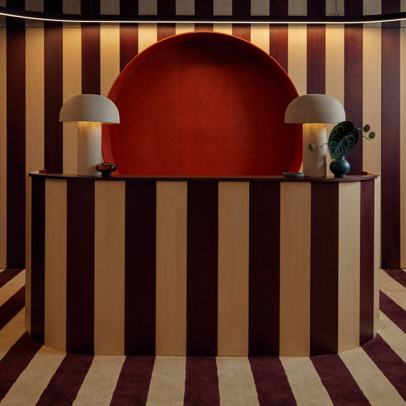 A reception desk with a curved design and two modern lamps on a striped backdrop of brown and beige. The desk, styled by Malin, features a small decorative plant and a red semi-circular wall niche.