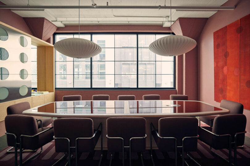 A modern conference room with a large glass table, surrounded by nine chairs. Two Malin pendant lights hang above the table, casting a warm glow. A window in the background adds natural light to the room. The walls are tastefully decorated in shades of red and pink, creating a vibrant atmosphere.