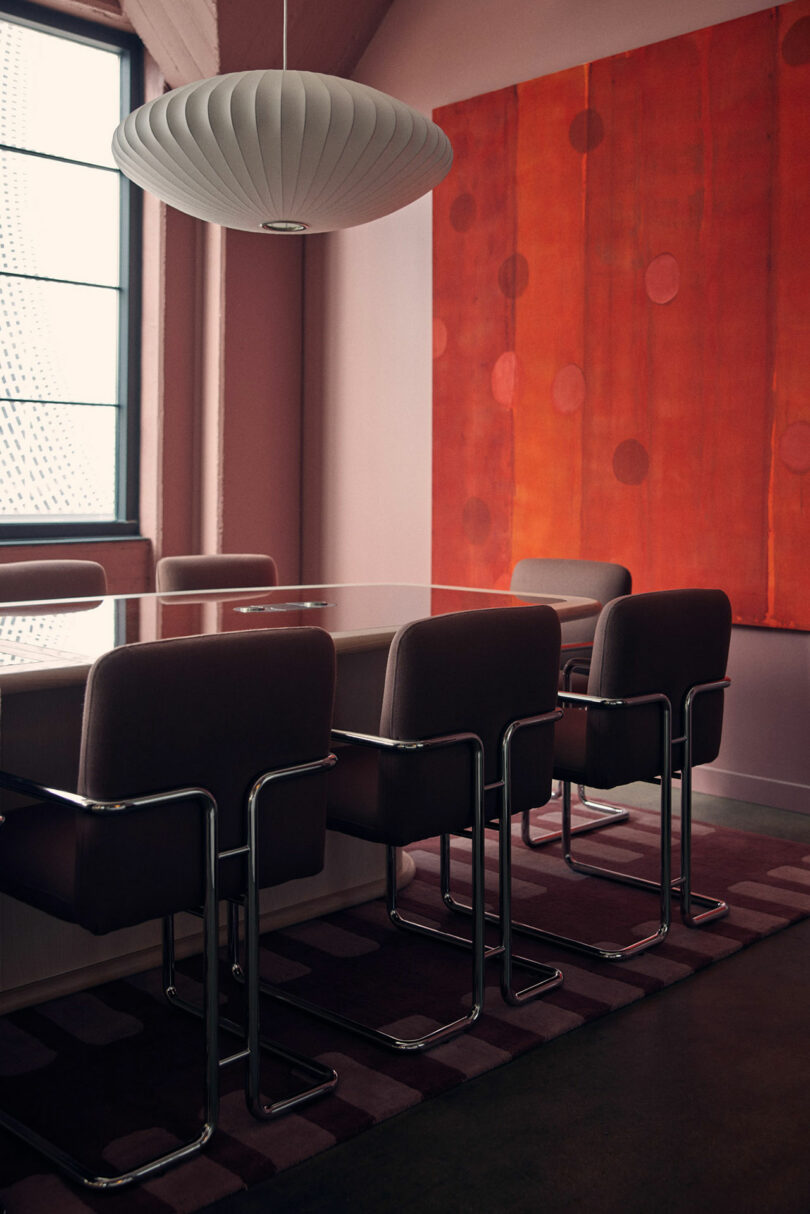 A modern conference room features a large table surrounded by chairs. A geometric red painting by Malin is mounted on the wall, and a contemporary light fixture hangs overhead.