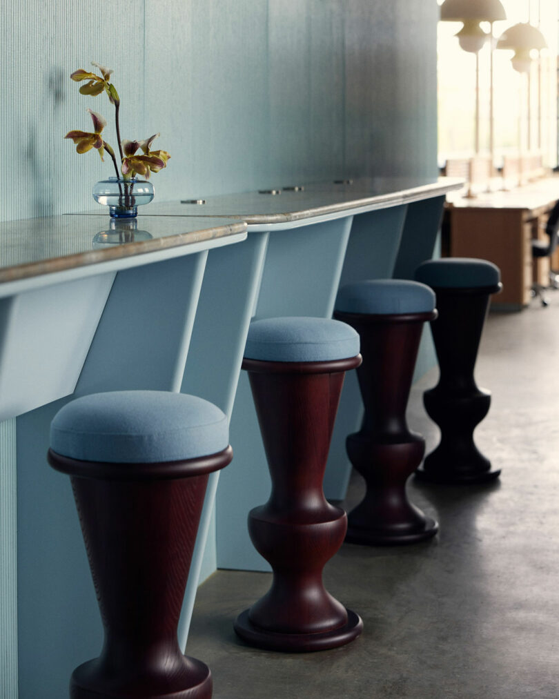 A sleek, modern bar features four tall, dark wood stools with Malin blue cushions, a light blue counter with minimalistic support, and a small vase with flowers.