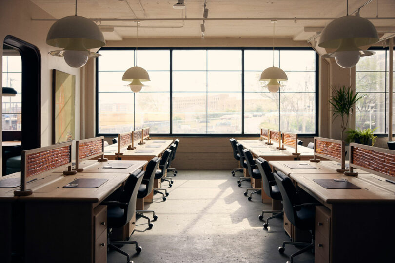 A well-lit, modern office with Malin wooden desks in rows, black chairs, partitions, large windows, plants, and hanging lights.