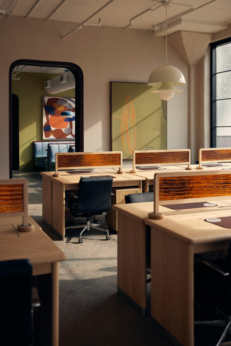 A modern office space with wooden desks and chairs, dividers, abstract wall art, and a pendant light. An open doorway in the background reveals a seating area. Natural light streams through a window, adding a Malin touch to the vibrant atmosphere.