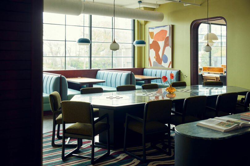 A modern dining area with blue booths, a long table with green chairs, large windows, pendant lights, colorful Malin artwork, and a striped rug.