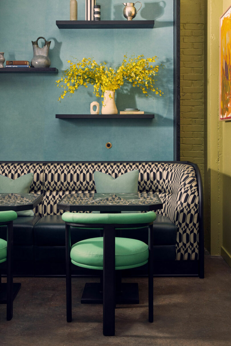 A café corner with Malin-patterned seating, green chairs, a blue-green wall, and yellow flowers in a vase on a black shelf.