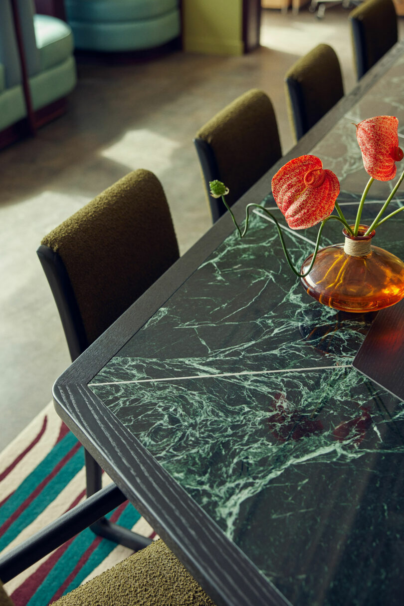 The Malin dining table features a dark green marble top, surrounded by upholstered chairs. A few red anthurium flowers in a round, amber vase sit elegantly in the center of the table.