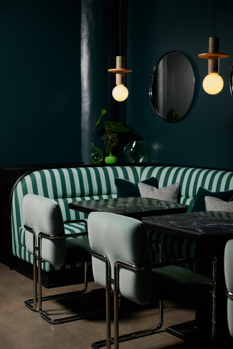 A modern dining area with a green and white striped Malin banquette, cushioned chairs, marble-topped tables, round mirrors, and hanging pendant lights.