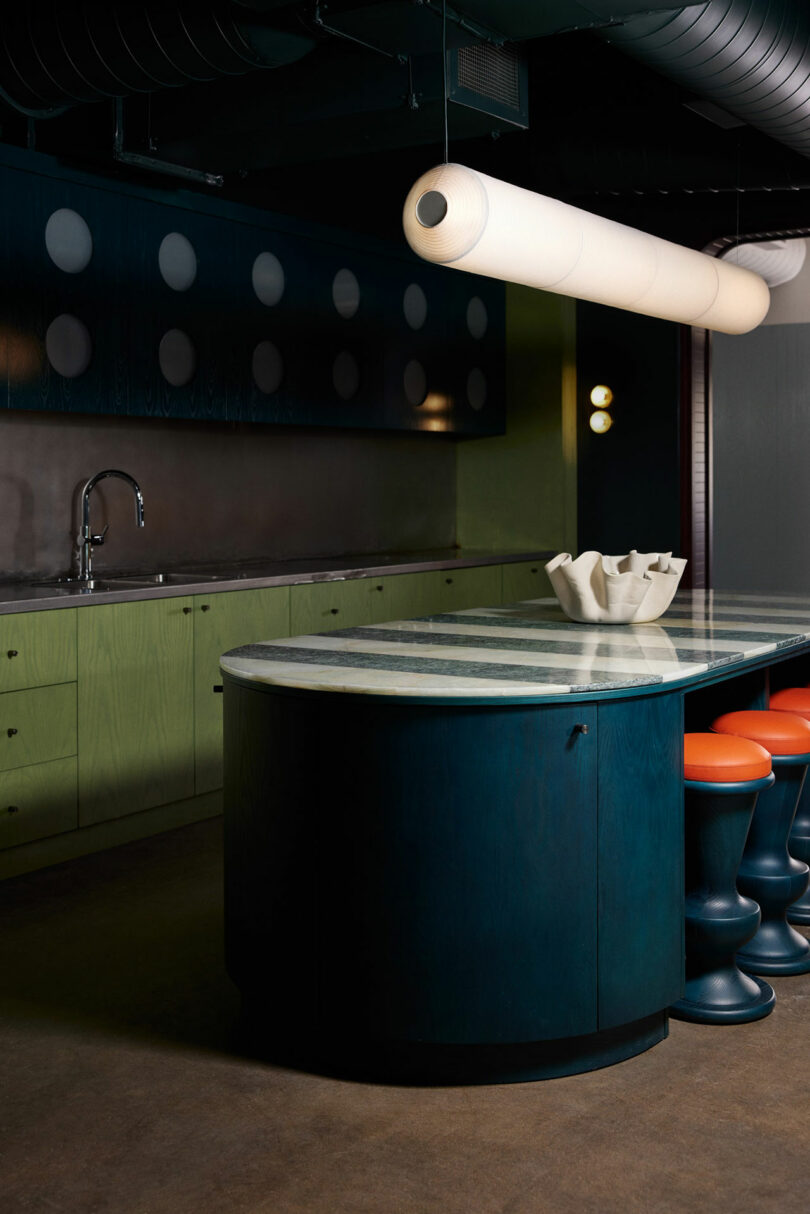 A modern kitchen features green cabinets, a curved island with a marble countertop, three orange Malin stools, a cylindrical light fixture, and a faucet on the countertop.