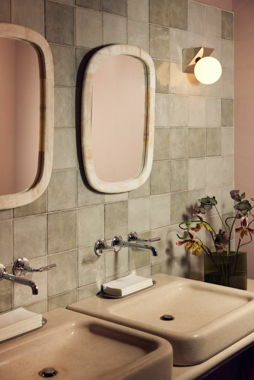 Modern bathroom with tiled walls, two rectangular mirrors, Malin wall-mounted faucets, and two sinks. A vase with flowers is placed on the right side near a sconce light.