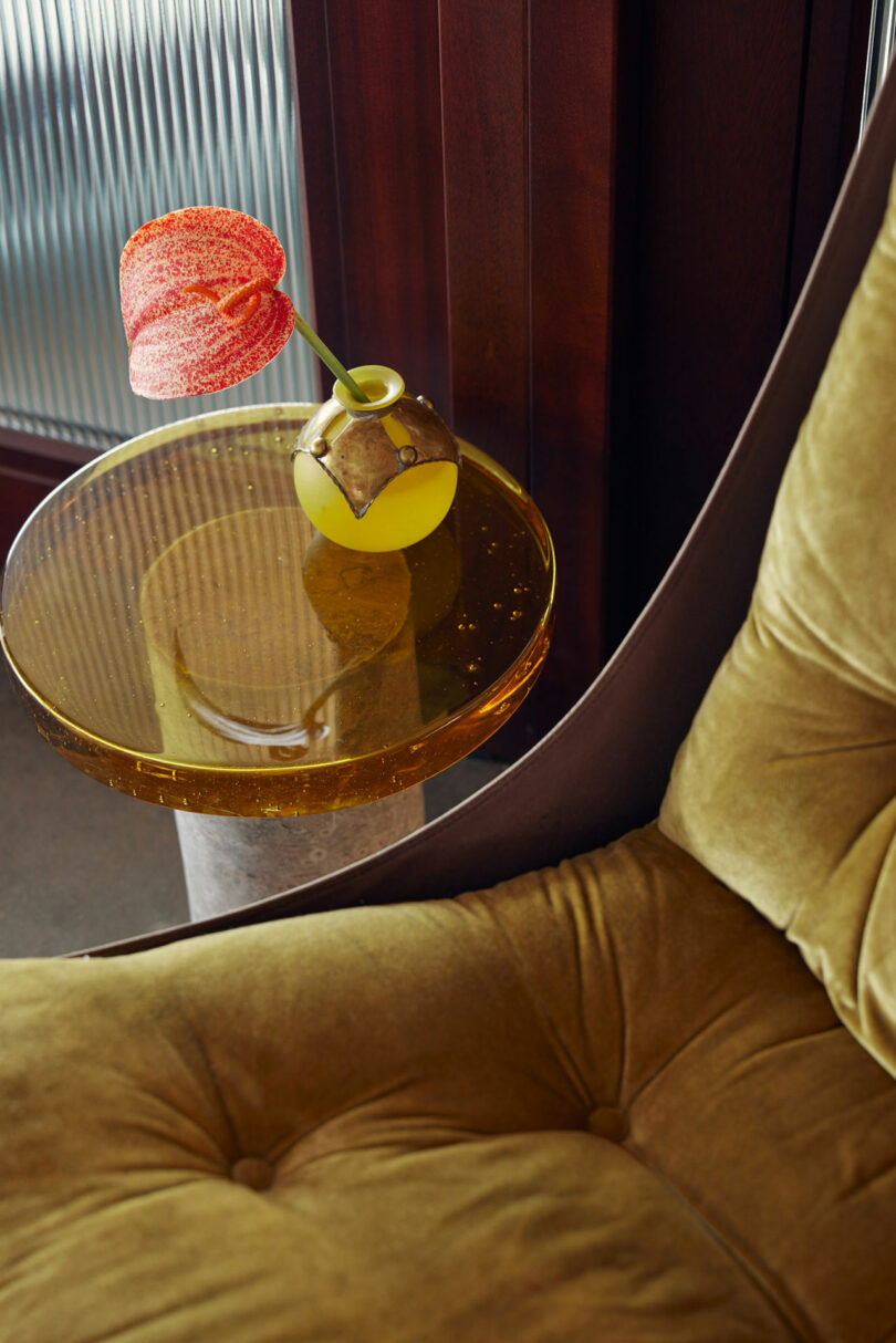 A small yellow vase with a single red flower on a round glass side table, next to a mustard-colored cushioned chair.