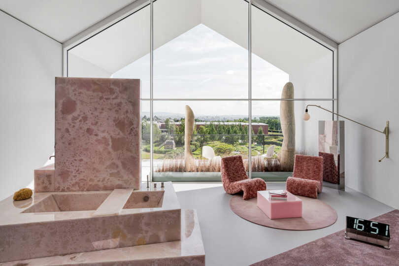 A modern room with a large window, pink stone furniture, two pink textured chairs, a pink rug, a clock showing 16:51, and a view of a landscaped garden and sculptures outside.
