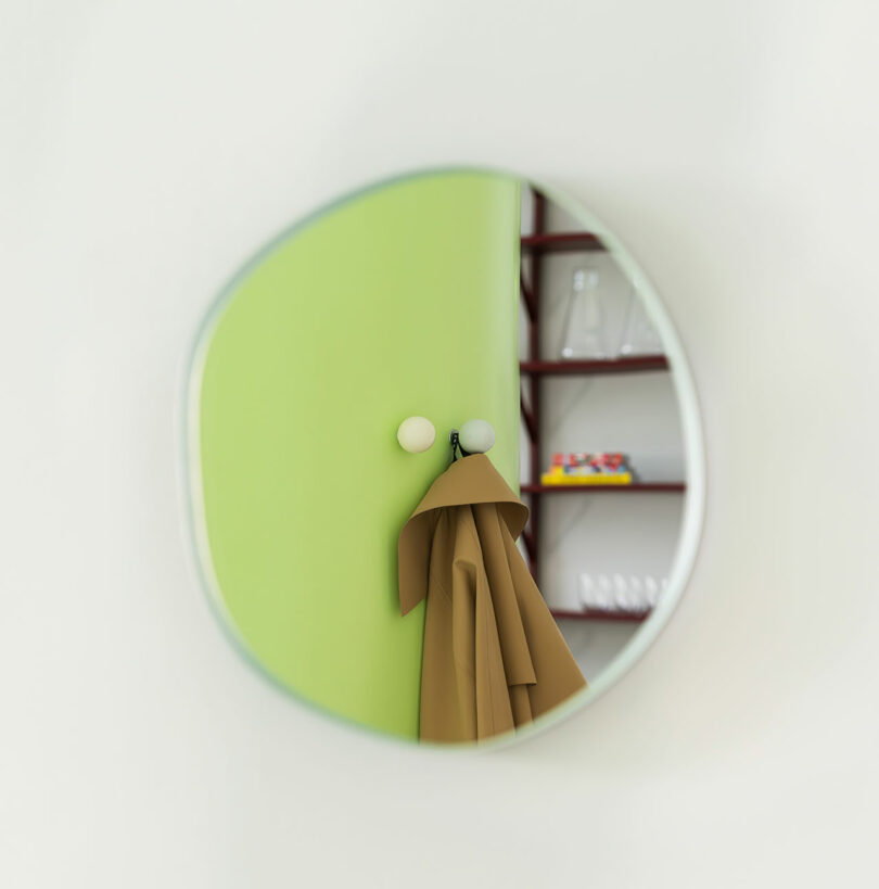 A round mirror reflects a green wall with two hooks, one holding a light brown coat. A shelf with glassware and colorful items is visible in the background.