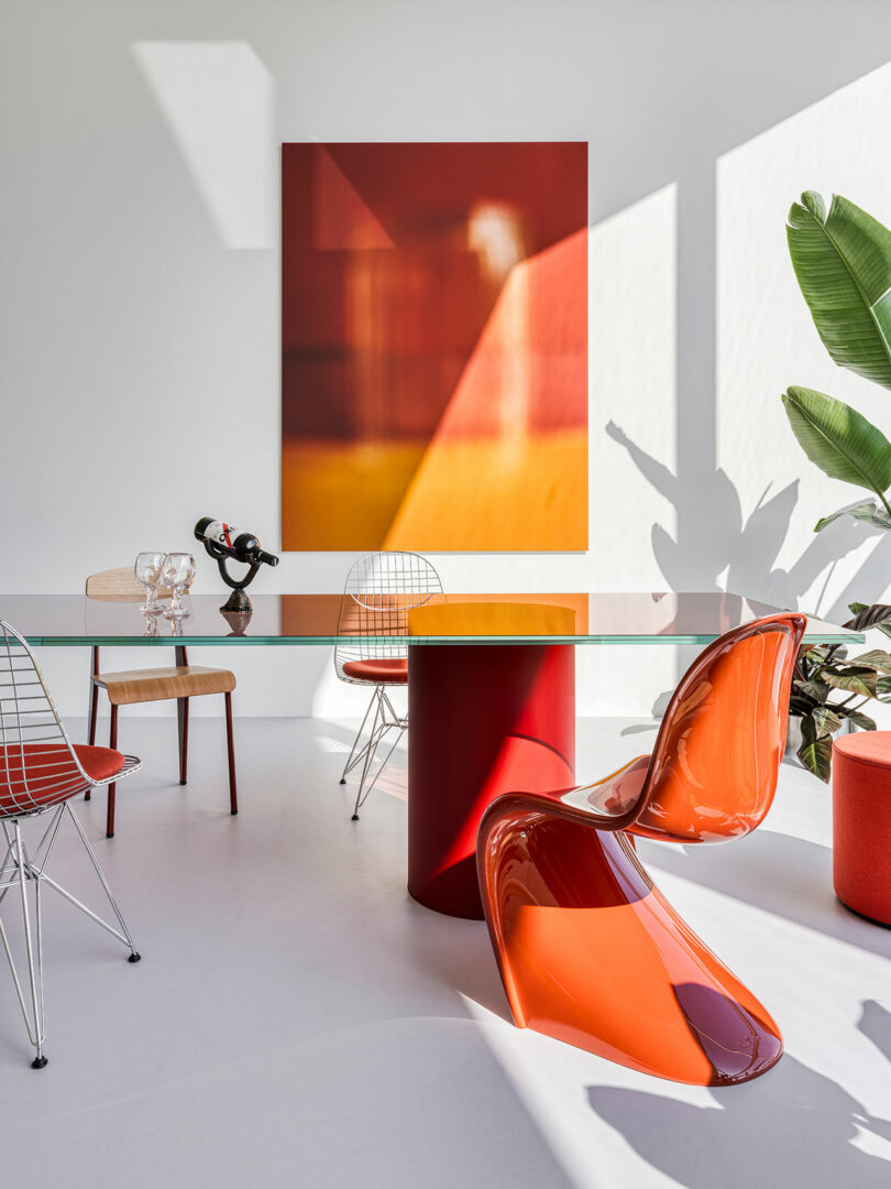 Modern dining room with a glass table, red cylindrical base, two red chairs, two wire chairs, abstract wall art, and a potted plant. Sunlight casts shadows on the wall and floor.